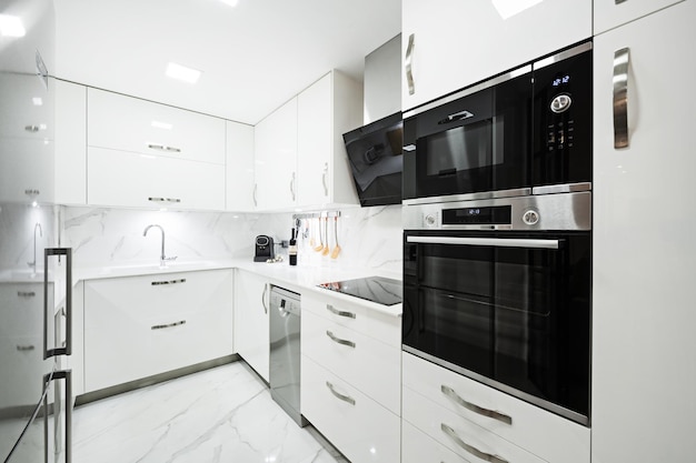 Foto cocina amoblada con muebles color blanco brillante sobres blancos lisos pisos y paredes de losetas de mármol blanco con vetas y electrodomésticos de acero inoxidable y vidrio negro