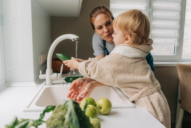 En la cocina brillante madre e hija pequeña lavan verduras para preparar la cena juntos