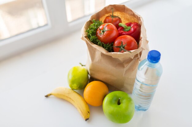 cocina, dieta, comida vegetariana y concepto de alimentación saludable: cierre de una bolsa de papel con frutas y verduras frescas y jugosas y una botella de agua en la mesa de la cocina en casa