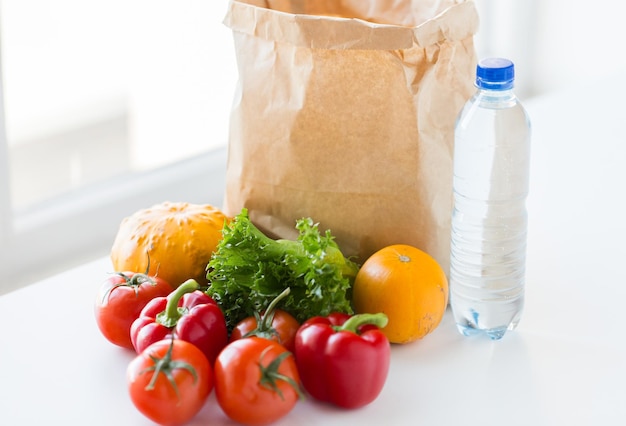 cocina, dieta, comida vegetariana y concepto de alimentación saludable: cierre de una bolsa de papel con verduras jugosas maduras frescas y una botella de agua en la mesa de la cocina en casa