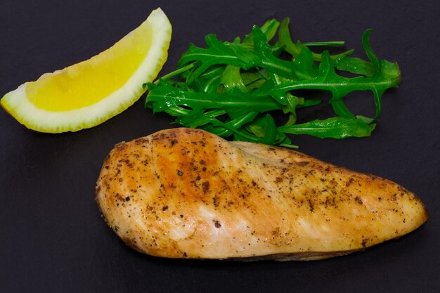 Cocina dietética: pechuga de pollo a la parrilla con hojas de rúcula y limón sobre fondo oscuro.