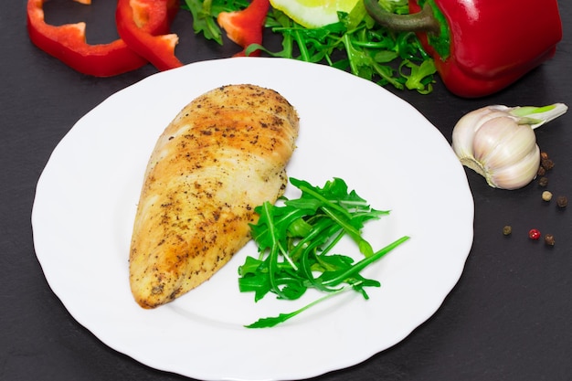 Foto cocina dietética: pechuga de pollo a la parrilla con hojas de rúcula y verduras de fondo oscuro.