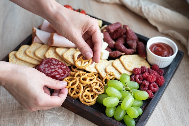 Cocina de fiesta, manos femeninas poniendo el mini pretzel en tablero de charcutería con salchicha, fruta, galletas y queso.