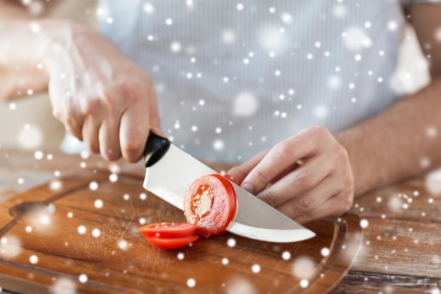 Foto cocina, gente, comida vegetariana y concepto de hogar - cerca de un hombre cortando tomate y otras verduras en una tabla de cortar con un cuchillo grande