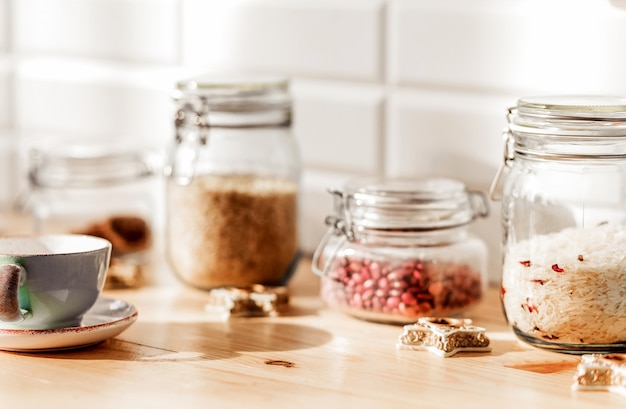 En la cocina hay frascos de arroz y frijoles. Junto a las latas hay una taza de té en un platillo y galletas. Foto horizontal