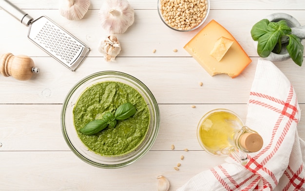 Cocina italiana. Preparación de salsa pesto italiana casera. Pesto fresco en un tazón con ingredientes, vista superior plana yacía sobre mesa de madera blanca