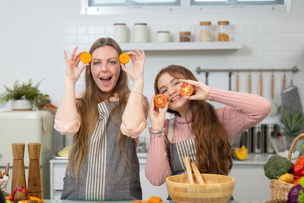 En una cocina moderna, blanca e impecable, una madre y una hija sonrientes demuestran cómo preparar verduras, tomates, juegan de cerca para la cena