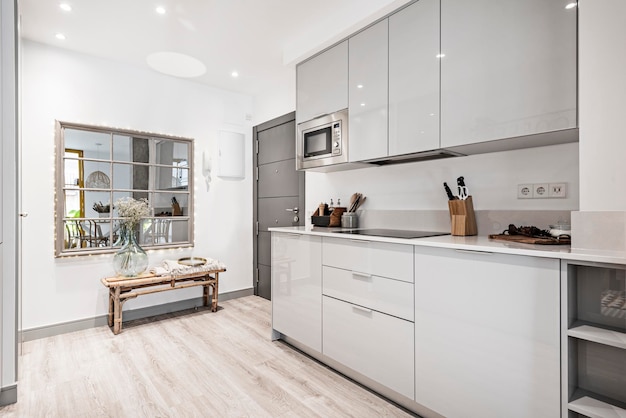 Cocina con muebles de madera gris combinados con encimera de piedra blanca  y armarios de pequeños electrodomésticos vitrocerámica y horno microondas  junto a la puerta de entrada a un apartamento de alquiler