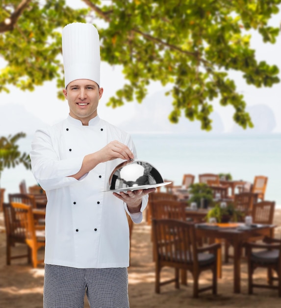 cocina, profesión y concepto de la gente - feliz chef masculino sosteniendo cloche sobre el salón del restaurante en la playa