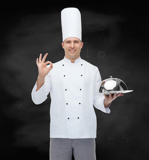 cocina, profesión, gesto y concepto de la gente - feliz chef masculino sosteniendo un cloche y mostrando un cartel de ok sobre el fondo de la pizarra negra