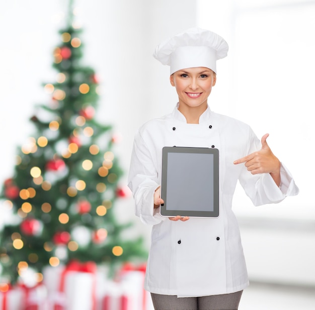 Cocina, vacaciones, tecnología y concepto de comida: chef, cocinera o panadera sonriente con una computadora de tableta apuntando con el dedo a la pantalla en blanco sobre la sala de estar con fondo de árbol de Navidad