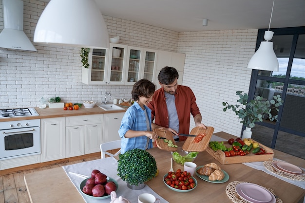 Cocinar el almuerzo con papá, padre joven y su hijo feliz preparando una ensalada mientras está de pie en el