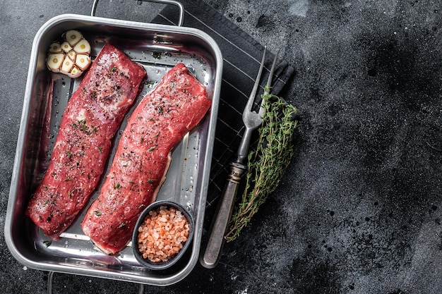 Cocinar la carne de solomillo de cordero con hierbas y aceite de oliva, carne de cordero marinada en bandeja de cocina. Fondo negro. Vista superior. Copie el espacio.