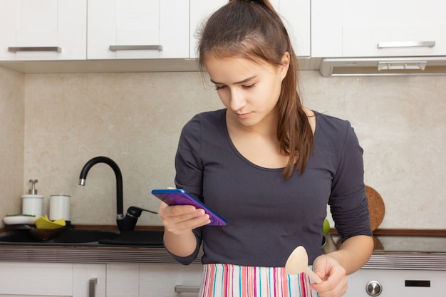 Cocinar comida deliciosa. Niña lee una receta en un teléfono inteligente en la cocina.