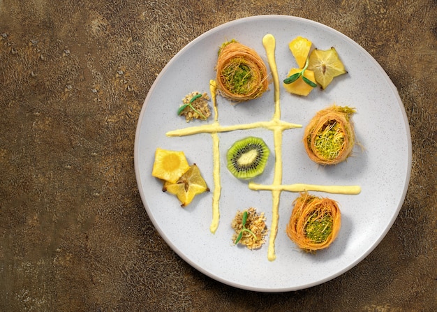 Cocinar dulces postres de pastelería de ramadan tradicional turco kunafa (kadaif, baklava), kiwi, ananas, nueces, fondo oscuro Composición de arte de placa.