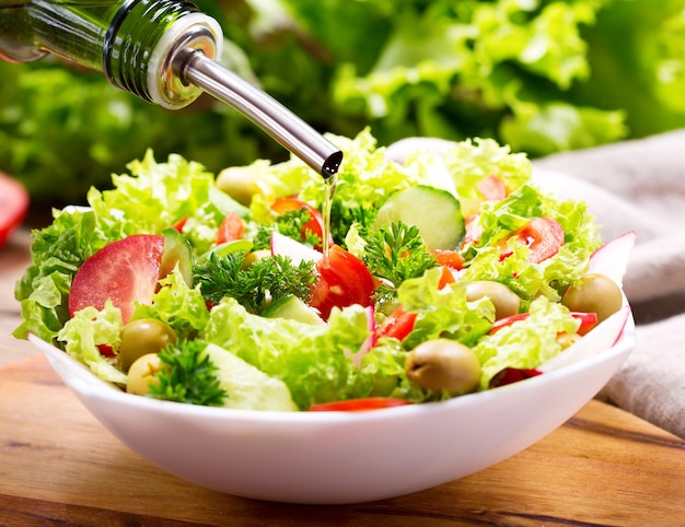 Cocinar ensalada de aceite de oliva verter en un tazón de ensalada fresca con verduras y verduras en la mesa de madera