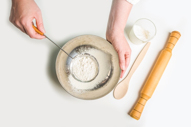 Cocinar la masa para hornear en la cocina de casa. Las manos femeninas sostienen un vaso de harina y un tamiz. Concepto de comida casera.