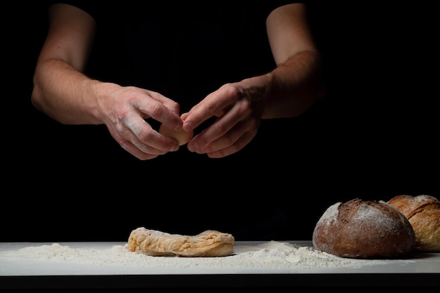 Cocinar el pan. Las manos del chef agregan un huevo de gallina que vuela sobre la mesa blanca con la harina y la masa. El chef espolvorea polvo de harina blanca con la mano de un hombre sobre un fondo negro. Espacio para texto.