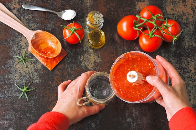 Cocinar salsa de tomate casera. Tomates y especias. Las manos femeninas en mangas largas rojas vierten salsa de tomate recién preparada de una licuadora en un frasco de vidrio.