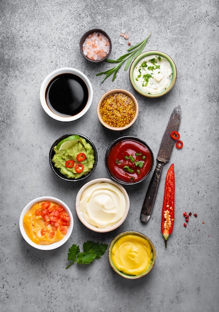 Cocinar salsas y aderezos saludables concepto. Diferentes salsas en cuencos sobre fondo de hormigón rústico, ingredientes, hierbas, ají picante y cuchillo de cocina, vista superior, primer plano