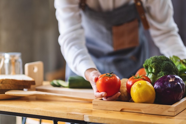 Una cocinera sosteniendo y recogiendo un tomate fresco de una bandeja de verduras en la mesa