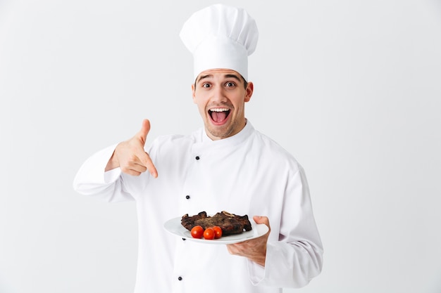 Cocinero alegre vistiendo pimientos uniformes carne de bistec cocida en un plato aislado sobre la pared blanca