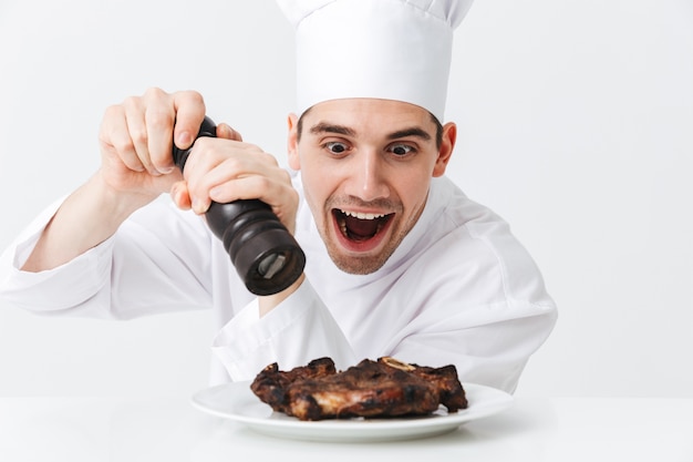 Cocinero alegre vistiendo pimientos uniformes filete de ternera cocida en un plato aislado sobre la pared blanca