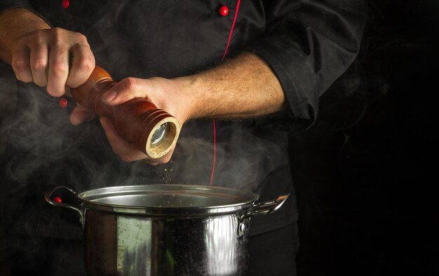 El cocinero añade pimienta molida a una olla de comida hirviendo Concepto de cocina de restaurante con espacio publicitario sobre fondo negro