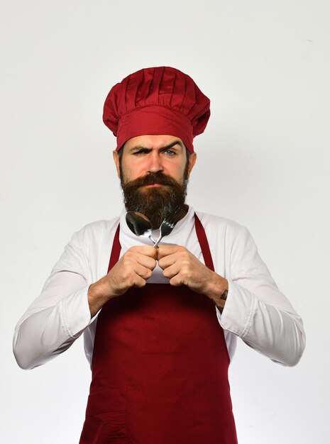 Foto cocinero con cara estricta en uniforme burdeos muestra cubiertos. concepto de catering de restaurante. chef cruza utensilios de cocina. el hombre con barba tiene tenedor y cuchara sobre fondo blanco.