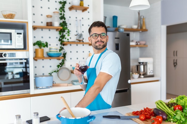 Cocinero casero Hombre soltero en delantal mirando la ventana bebiendo vino de un vaso mientras cocina en la cocina cocinando en casa