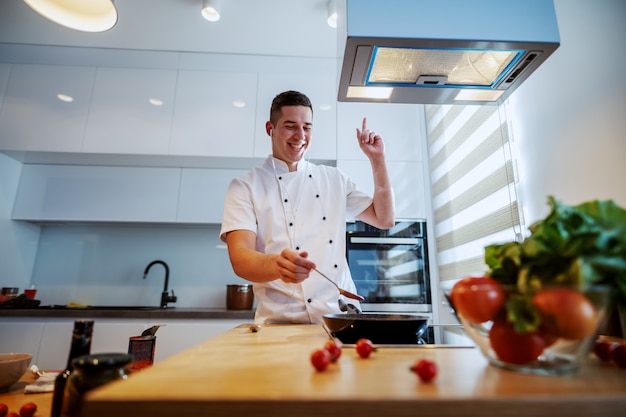 Cocinero caucásico alegre en uniforme disfrutando de la música y preparando la salsa de tomate mientras está de pie en la cocina.