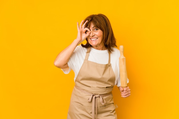 Cocinero caucásico de mediana edad mujer sosteniendo un rodillo aislado emocionado manteniendo el gesto ok en el ojo.