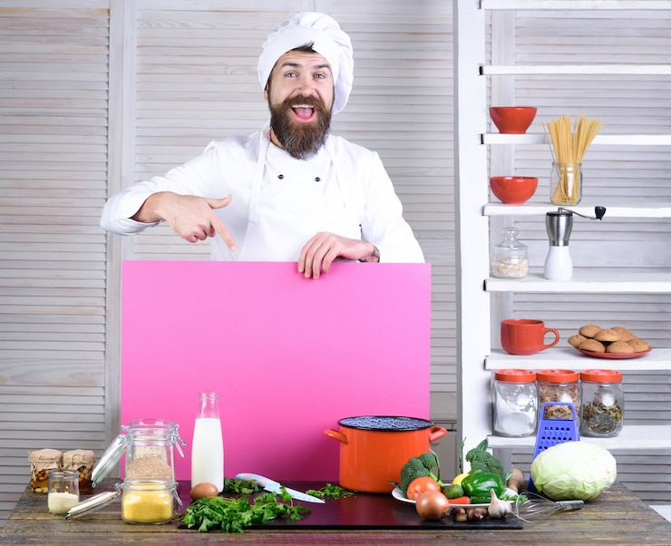 Foto cocinero de chef barbudo feliz sostiene tablero rosa para publicitar chef profesional en servicio de comida de cocina