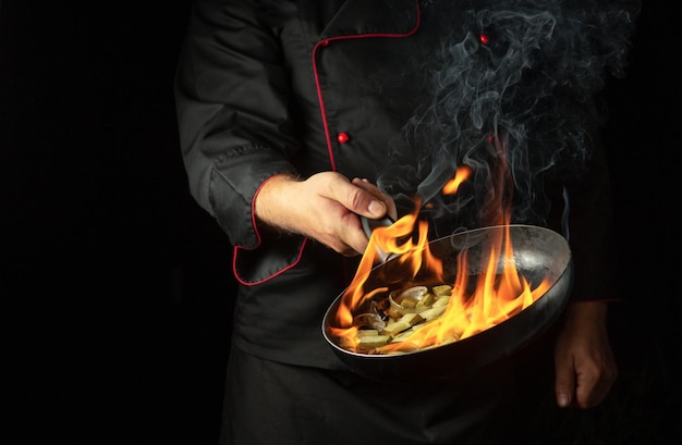 El cocinero cocinando comida en una sartén con la llama del fuego del ron El concepto de cocina del restaurante en fondo negro con espacio para el menú o la receta Flambler o cocinar verduras en fuego natural