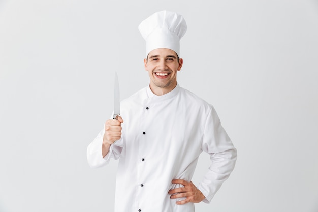 Cocinero cocinero feliz vistiendo uniforme sosteniendo un cuchillo aislado sobre la pared blanca