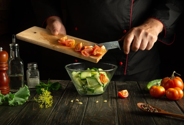 El cocinero está preparando una ensalada de verduras en la cocina El concepto de cocinar ensalada de vitaminas con tomates y pepinos Espacio para una receta en un fondo oscuro