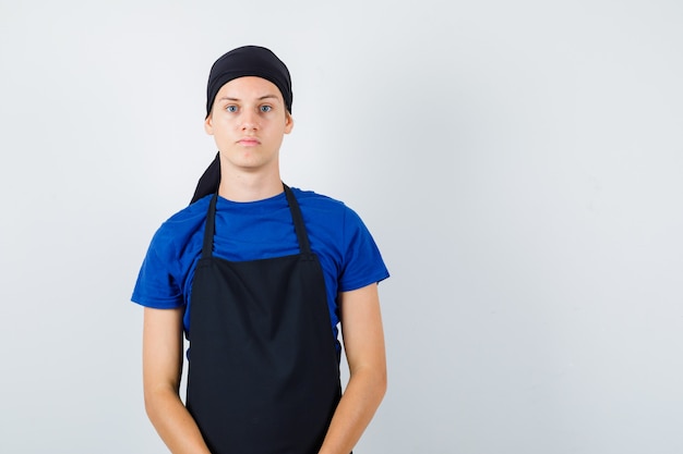 Cocinero joven mirando al frente en camiseta, delantal y mirando pensativo. vista frontal.