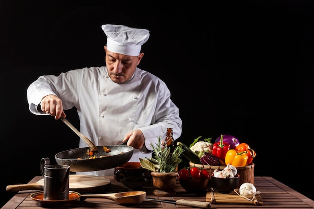 Cocinero masculino con uniforme blanco y sombrero con cucharón mezcla los ingredientes en la sartén