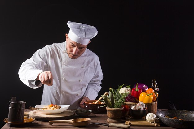 Cocinero masculino en uniforme blanco y sombrero poniendo hierbas en un plato de comida con vegetales