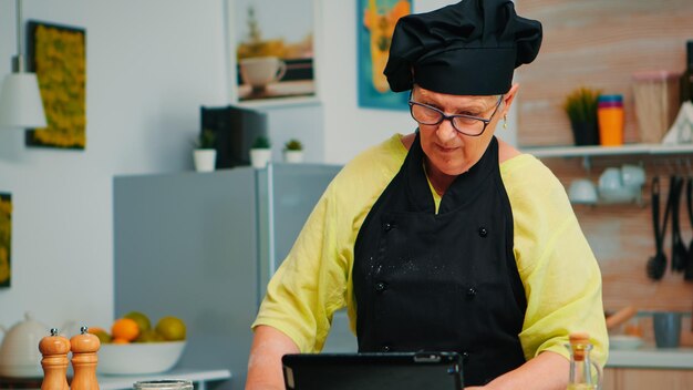 Cocinero de la mujer que usa la tableta en la cocina mientras se cocina la pizza. Señora jubilada siguiendo un consejo culinario en una computadora portátil, aprendiendo un tutorial de cocina en las redes sociales, usando un rodillo de madera para formar la masa.