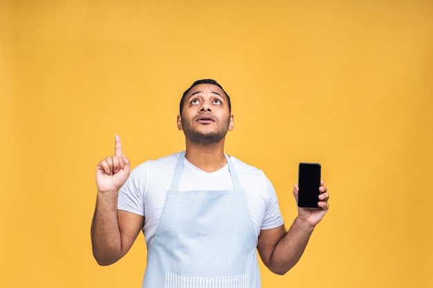 Cocinero o chef indio afroamericano masculino negro tocando la pantalla del teléfono móvil y apuntando con el dedo índice a la pantalla en blanco, aislada sobre fondo amarillo, espacio de copia, recorte. Dedo señalador.