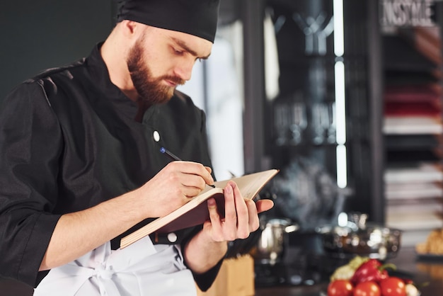 Cocinero profesional joven en uniforme de pie con bloc de notas en la cocina