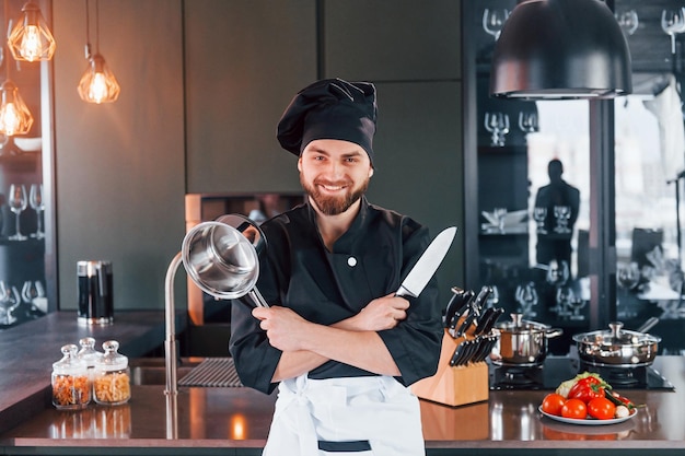 Cocinero profesional joven en uniforme de pie cerca de la mesa y posando para una cámara en la cocina