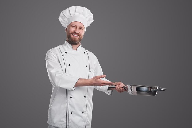 Cocinero profesional masculino en uniforme de chef blanco demostrando una sartén de acero inoxidable y sonriendo amigablemente mientras recomienda utensilios de cocina contra el fondo gris