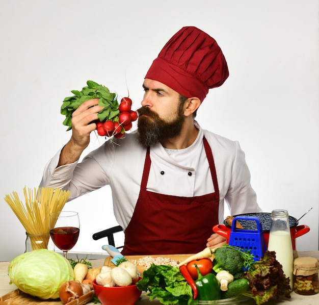 Cocinero con rostro serio en uniforme burdeos sentado junto a la mesa de la cocina con verduras y utensilios de cocina Concepto de proceso de cocción Hombre con barba huele a rábano sobre fondo blanco El chef prepara la comida