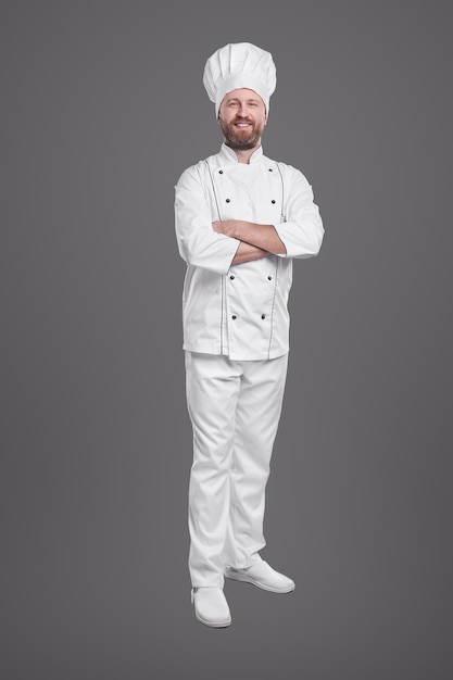 Cocinero de sexo masculino alegre vistiendo uniforme blanco y gorra de pie con los brazos cruzados sobre fondo gris en estudio