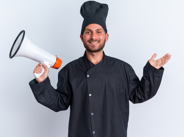 Cocinero de sexo masculino caucásico joven alegre en uniforme del cocinero y la tapa que sostiene el altavoz que muestra la mano vacía