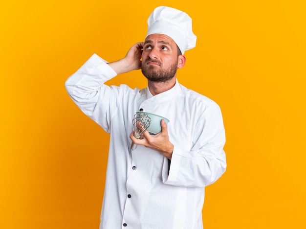 Cocinero de sexo masculino caucásico joven confuso en uniforme del cocinero y la tapa que sostiene el batidor y el tazón de fuente que mira hacia arriba rascándose la cabeza aislada en la pared naranja
