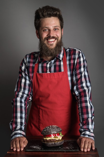 Cocinero de sexo masculino en delantal rojo de pie cerca de una hamburguesa negra fresca hecha a sí misma y sonriendo