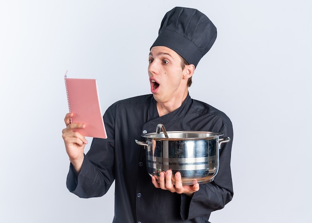 Cocinero de sexo masculino rubio joven sorprendido en uniforme del cocinero y el casquillo que sostiene el bloc de notas de lectura de la olla aislado en la pared blanca